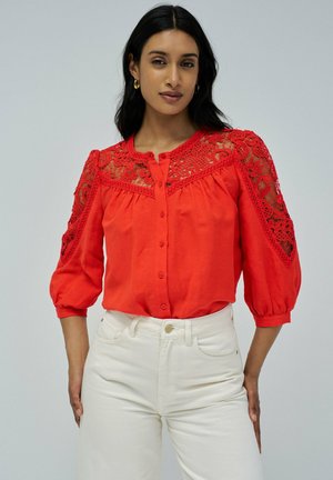 Blusa roja con mangas largas y abullonadas, con detalles de encaje en los hombros y un cierre frontal de botones. Combinada con jeans blancos.
