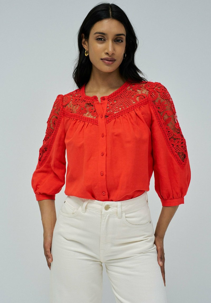 Blusa roja con mangas largas y abullonadas, con detalles de encaje en los hombros y un cierre frontal de botones. Combinada con jeans blancos.