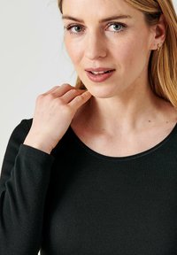 Maglietta nera a maniche lunghe con collo rotondo. Il modello ha una carnagione chiara, con morbide onde nei capelli biondi e sta toccando il collo.