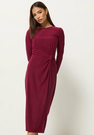 Femme aux longs cheveux foncés portant une robe bordeaux ajustée à manches longues avec un détail de nœud froncé à la taille, debout devant un fond uni.