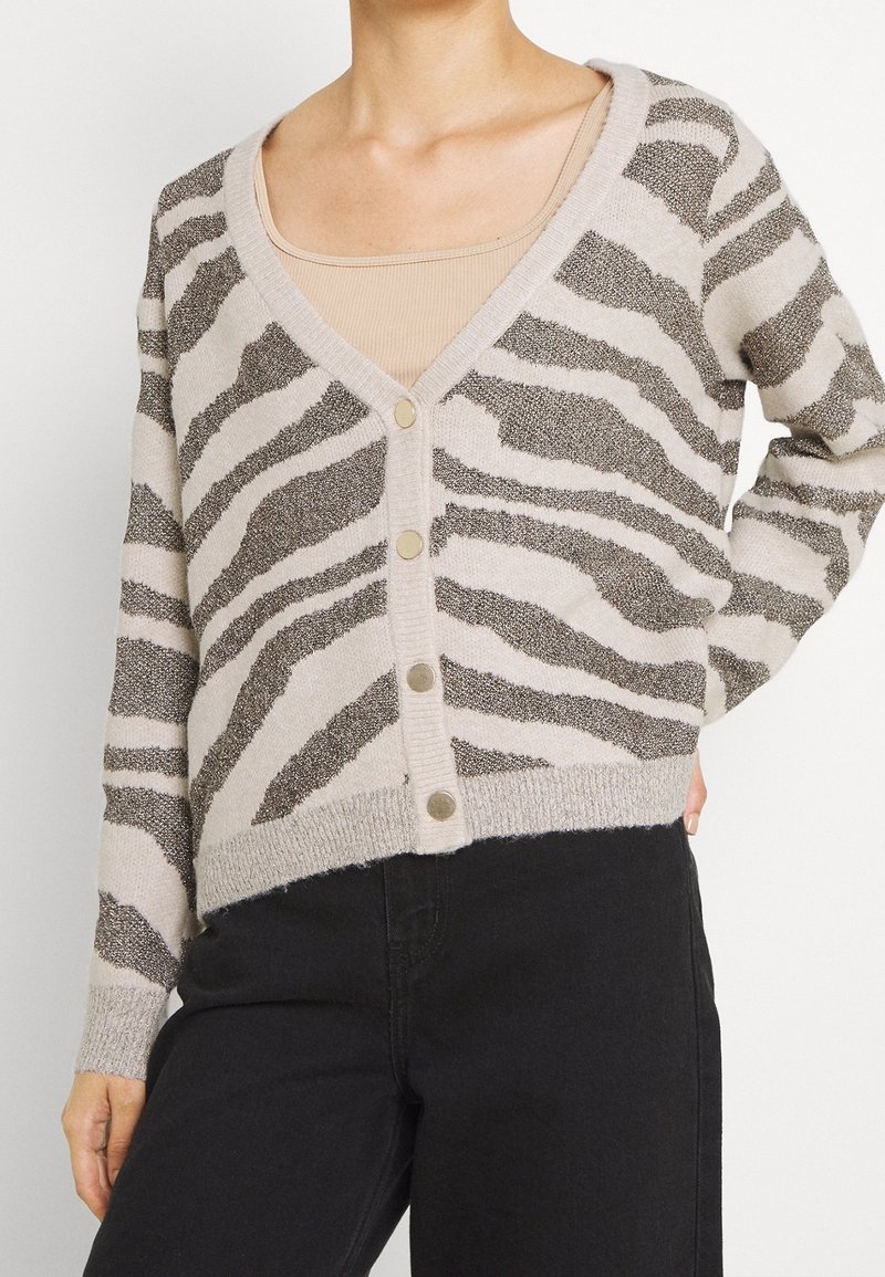 Femme portant un cardigan boutonné beige et marron à rayures zébrées sur un top beige avec un pantalon noir, main posée sur la hanche.