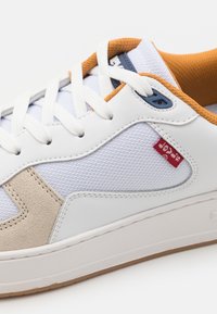 Hvide sneakers med en kombination af læder og mesh i overdelen. Funktioner inkluderer beige accenter, orange krave, hvide snørebånd og et lille rødt mærke på siden.