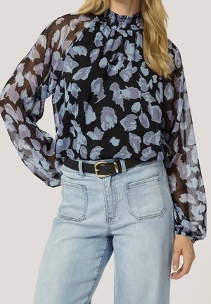 Zwarte semi-transparante blouse met blauw- en paarse bloemenprint, lange pofmouwen, hoge geplooide hals, ingestopt in lichtblauwe spijkerbroek met zwarte riem.