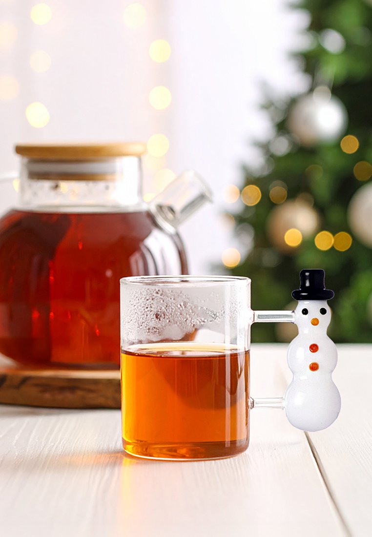 Przezroczysty szklany kubek wypełniony bursztynowym płynem, z uchwytem w kształcie bałwanka, który ma czarny kapelusz i pomarańczowe guziki, ustawiony obok imbryka.