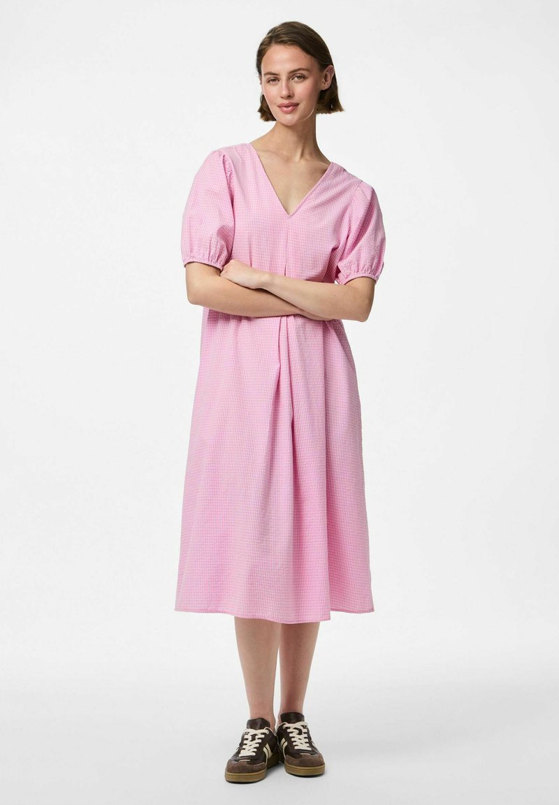 Roza karo midi haljina s napučenim rukavima, V-izrezom i nabranim strukom, u kombinaciji s tamnosmešnim patikama koje imaju bele vezice.