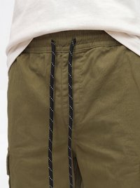 Gros plan d'un pantalon vert olive avec des cordons noirs à motif à pois blancs, porté avec une chemise beige.