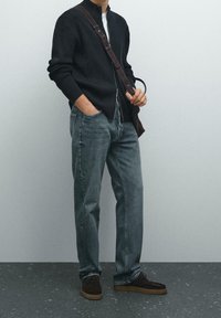 Mies, joka on pukeutunut mustaan vetoketjulliseen neuletakkiin, harmaisiin farkkuihin ja ruskeisiin kenkiin, kantaa tummaa ruskeaa olkalaukkua yksinkertaista taustaa vasten.