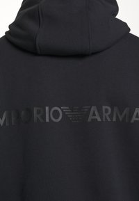Černá mikina s kapucí, vyrobená z texturované látky, s velkým matně černým logem "EMPORIO ARMANI" umístěným na zádech.