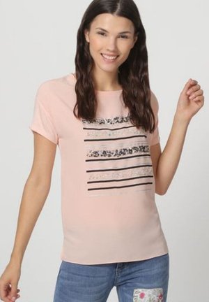 Joven con el pelo largo y oscuro que lleva una camiseta rosa con franjas de lentejuelas negras y plateadas y vaqueros azules con un parche floral plateado.