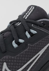 Chaussure de sport noire avec une tige texturée, des accents argentés et des lacets bleus. Dotée d'une languette rembourrée et d'un design épuré.