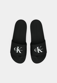 Black slide sandals with a smooth synthetic strap. Features white "Calvin Klein" and "CK" logos. Textured footbed for grip.