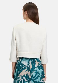 Biała teksturowana skórzana kurtka o długości do bioder z rękawami trzy czwarte. Bez widocznych zapięć. Zestawiona z plisowaną spódnicą w kwiatowy wzór.