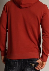 Röd hoodie med dragsko i huvan, mjukt tyg, ribbade ärmslut och nederkant; bärs med mörka denimjeans som har en etikett på bakfickan.