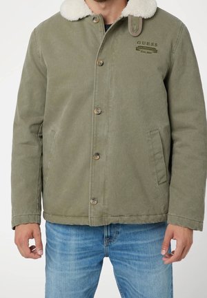 Hombre con chaqueta verde oliva abotonada con cuello de borreguito blanco y bolsillos delanteros, combinado con vaqueros azul claro.