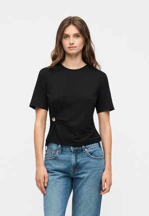 Young woman with wavy brown hair wearing a black short-sleeve top with a side button detail and blue denim jeans, standing straight.