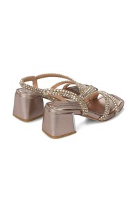 Sandalias de color oro rosa metálico con tacones cuadrados y gruesos, que presentan correas cruzadas adornadas con pequeños strass. Textura suave y parte trasera cerrada.