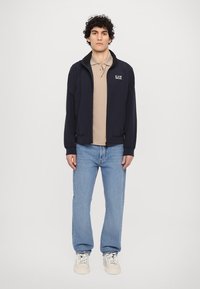 Cazadora de marina con cremallera frontal, puños elásticos y logo bordado. Usada sobre un polo beige, combinada con jeans azul claro y zapatillas blancas.