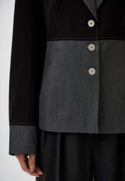 Black and gray blazer featuring button closure, contrasting stitched seams, and textured fabric. Long sleeves with gray cuff detail.