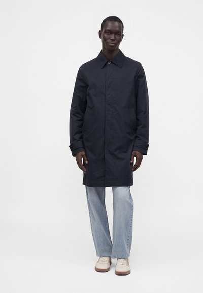 Homme debout portant un manteau noir mi-long, un jean bleu clair et des baskets blanches sur un fond clair uni.