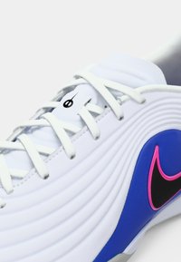 Λευκό αθλητικό παπούτσι με υφή στην επιφάνεια, με μπλε και ροζ λεπτομέρειες, στρογγυλεμένο σχήμα και επίπεδα λευκά κορδόνια. Μαύρο λογότυπο στο πλάι.