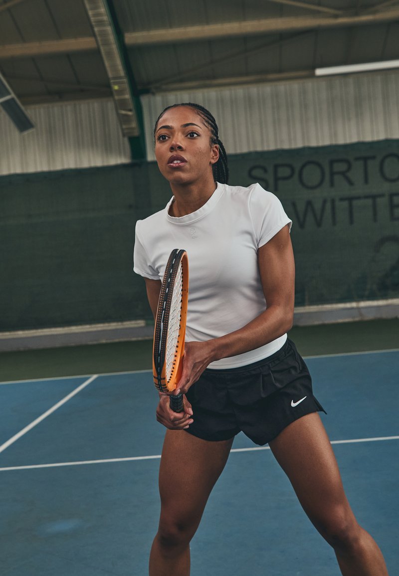 White athletic top and black shorts, holding an orange tennis racket, on a blue court with a green backdrop. Athletic build, dynamic pose.
