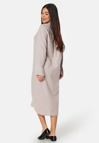 Camicia lunga a righe marrone chiaro e bianca, con maniche lunghe e una vestibilità rilassata. Sul retro presenta una leggera apertura per facilitare i movimenti. Abbinata a ballerine nere.