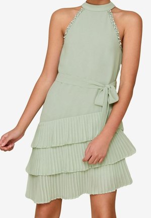 Robe sans manches vert pâle avec col halter, bretelles ornées, ceinture à la taille et jupe plissée en couches portée par une personne les bras détendus.