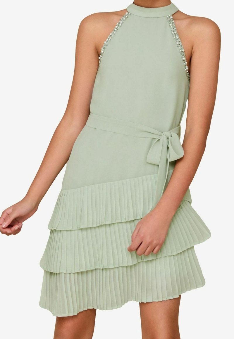 Robe sans manches vert pâle avec col halter, bretelles ornées, ceinture à la taille et jupe plissée en couches portée par une personne les bras détendus.