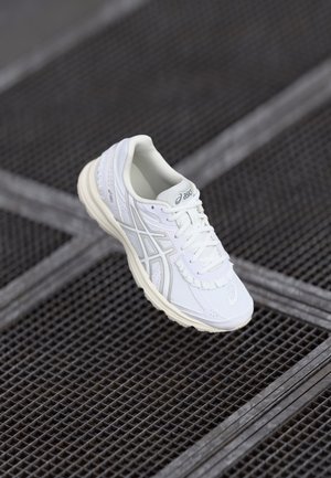 White athletic shoe with textured synthetic upper, curved sole, and tonal accents. Features laces and brand logo on the side.