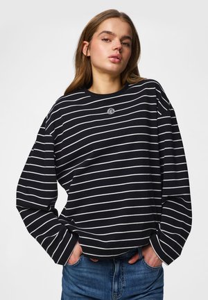 Mladá žena v čierno-bielom horizontálne pruhovanom tričku s dlhým rukávom a modrých džínsoch, ruky v vreckách, na jednofarebnom pozadí.