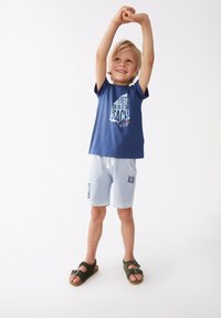 Camiseta azul con texto blanco y rojo, pantalones cortos azul claro y sandalias verdes. Niño posando con los brazos en alto, sonriendo. Diseño simple, estilo casual.