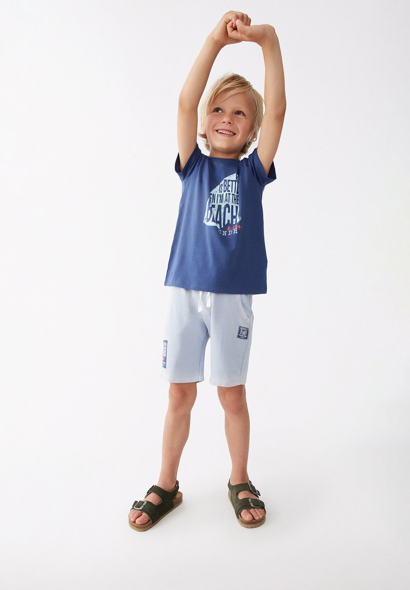 Camiseta azul con texto blanco y rojo, pantalones cortos azul claro y sandalias verdes. Niño posando con los brazos en alto, sonriendo. Diseño simple, estilo casual.