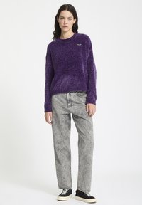 Lilla, struktureret striktrøje med runde hals og broderet logo, sat sammen med lysegrå, vide jeans og sorte sneakers.