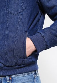 Giacca di denim blu scuro con polsini e orlo elasticizzati. Presenta tasche laterali e dettagli di cucitura in vista. Design casual.