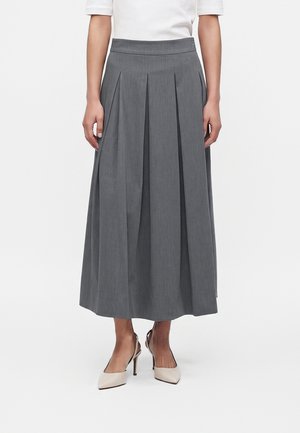 Frau trägt einen grauen plissierten Midirock und weiße Slingback-Pumps mit Absatz, steht vor einem einfarbigen weißen Hintergrund.