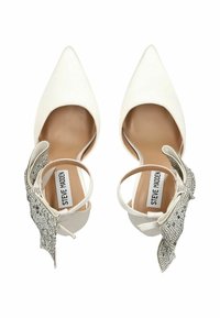 White pointed-toe high heels with ankle straps adorned with large rhinestone-studded bows, labeled Steve Madden inside.