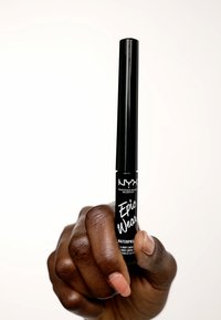 Eyeliner noir imperméable dans un tube cylindrique avec une étiquette blanche. Tenue dans une main avec un ongle rose clair court. Fond blanc uni.