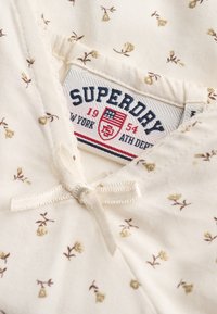 Gros plan d'un tissu crème avec de petits motifs floraux marron, avec une étiquette de la marque Superdry et un détail de nœud cousu.