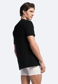 Black cotton T-shirt with short sleeves, straight cut, and a smooth texture. Worn with white fitted briefs, shown from the back.
