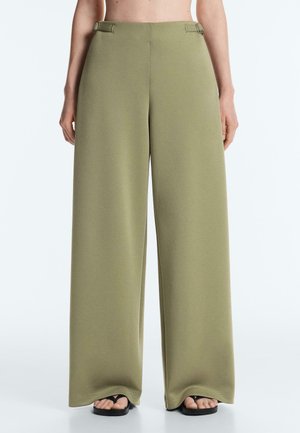 Szerokie spodnie w kolorze oliwkowym z regulowanymi klamrami po bokach, noszone z czarnymi sandałami z odkrytymi palcami.