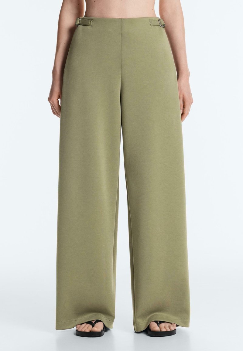 Pantalon large vert olive avec des boucles latérales réglables, porté avec des sandales noires à bouts ouverts.