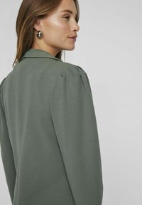 Olivově zelený blazer se strukturovaným límcem, nabíranými rameny a hladkým materiálem. Design zahrnuje upnutou siluetu a čisté linie.