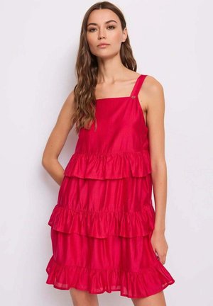 Donna dai capelli castano lunghi che indossa un abito rosso senza maniche a balze, in piedi davanti a uno sfondo bianco uniforme.