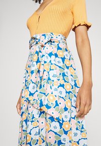 Haut orange à côtes sans manches associé à une jupe bleue à motifs floraux, ornée de fleurs roses, jaunes et vertes, nouée à la taille.