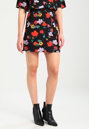 Femme portant une mini jupe noire avec un imprimé floral rouge, violet et jaune, associée à des bottes à talons pointus noires.