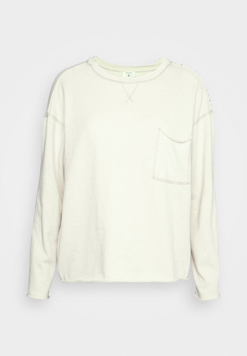 Sweat-shirt à manches longues couleur crème avec col ras du cou et poche poitrine, présentant des coutures apparentes et une coupe décontractée.