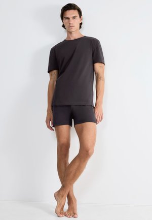 Mand står barfodet mod en hvid væg iført sort oversized t-shirt og sorte tætsiddende shorts, med synlige tatoveringer på arme og ben.