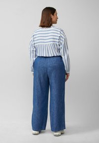 Gestreepte, lichte langslijfsblouse in wit en blauw, gecombineerd met losvallende blauwe spijkerbroek, gezien vanuit de achterkant.