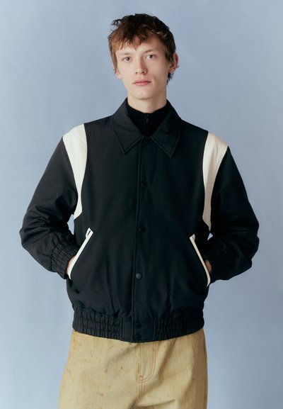 Black jacket with white accent panels, button closure, elasticized cuffs and waist, paired with light-colored pants. Soft texture.