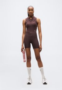 Γυναίκα με καφέ αμάνικο αθλητικό τοπ και σορτς που κρατάει ροζ ελαστική ζώνη, φοράει λευκές κάλτσες και αθλητικά παπούτσια, στέκεται μπροστά σε λευκό φόντο.